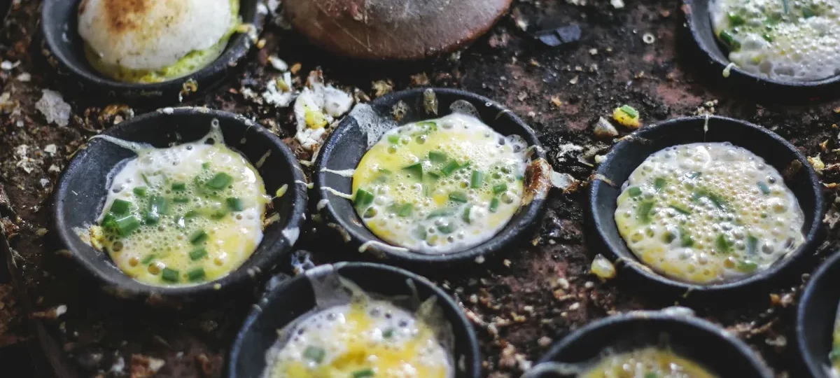 Bánh căn mini rice pancakes with quail egg and spring onion cooking in clay moulds at a Dalat street stall