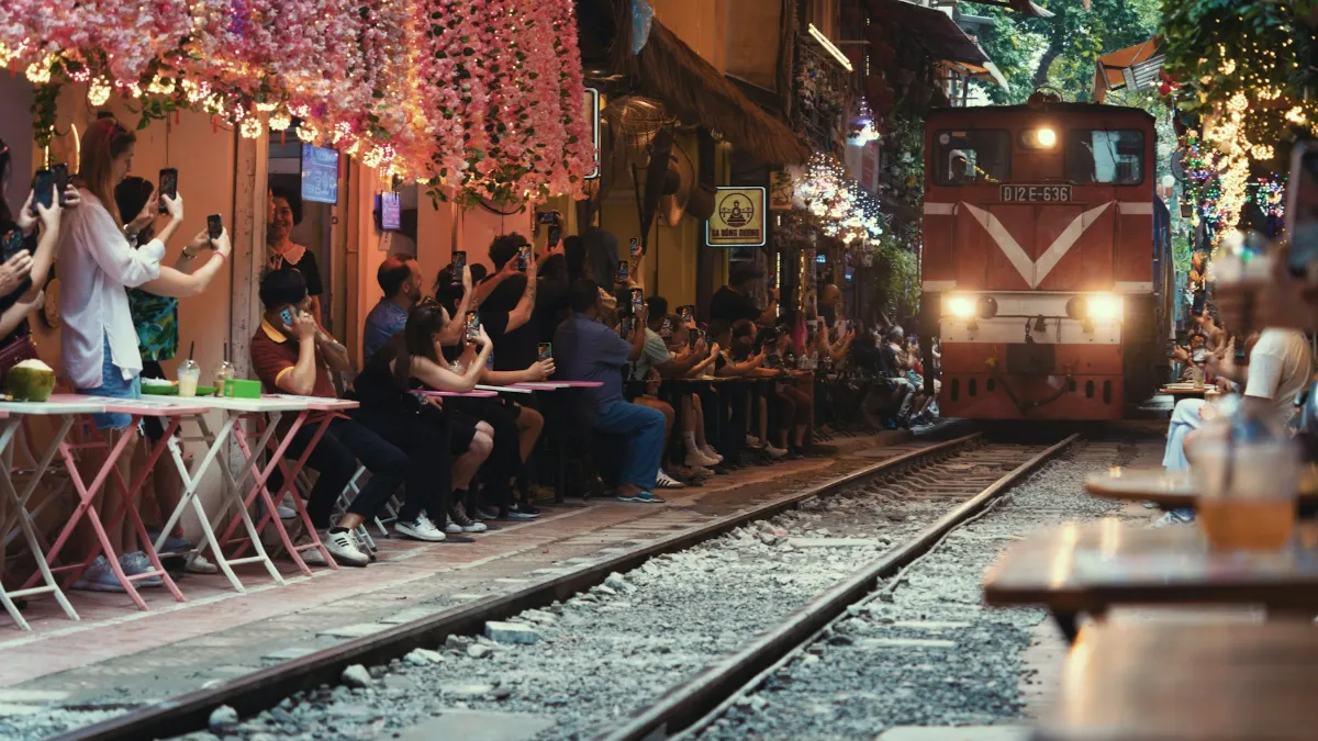 Hanoi Train Street – Train Approaching a Trackside Café A red locomotive approaches visitors seated at a trackside café on Hanoi Train Street
