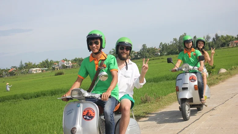 Guest and Vespa driver riding a country lane through rice fields near Hoi An